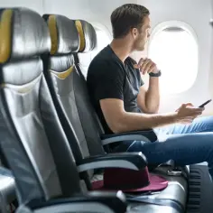 Mężczyzna w czarnej koszulce i jeansach siedzi przy oknie w samolocie, trzymając telefon. Zastanawia się, ile wcześniej być na lotnisku.